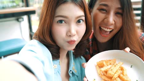 Beautiful-happy-Asian-women-lesbian-lgbt-couple-sitting-each-side-eating-a-plate-of-Italian-seafood-spaghetti-and-french-Fries-at-restaurant-or-cafe.-Female-couple-using-smartphone-for-selfie.