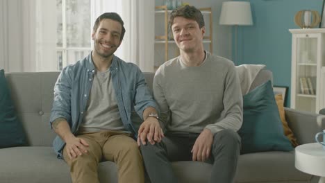 Cute-Attractive-Male-Gay-Couple-Sit-Together-on-a-Sofa-at-Home.-Boyfriend-Puts-His-Hand-on-Fiance's.-They-are-Happy-and-Smiling.-They-are-Casually-Dressed-and-Their-Room-Has-Modern-Interior.