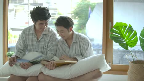 Young-asian-gay-couple-sitting-on-the-windowsill-and-reading-book-at-home-in-the-morning.