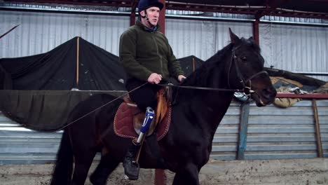 in-special-hangar,-a-young-disabled-man-learns-to-ride-a-black,-thoroughbred-horse,-hippotherapy.-man-has-an-artificial-limb-instead-of-his-right-leg.-concept-of-rehabilitation-of-disabled-with-animals