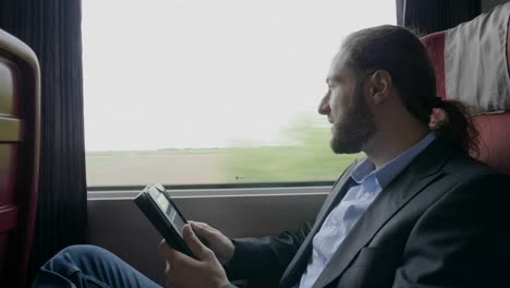 Happy-young-entrepreneur-man-commuting-on-train-relaxing-his-business-trip-watching-through-the-window-and-browsing-internet-on-tablet-pc