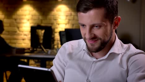 Closeup-portrait-of-young-cheerful-caucasian-businessman-typing-on-the-tablet-and-smiling-in-the-office-on-workplace