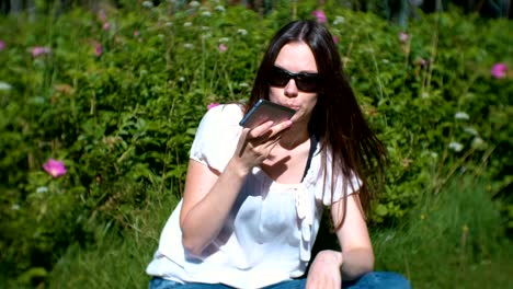 Beautiful-brunette-woman-records-a-voice-message-on-her-mobile-phone-while-sitting-in-the-park-on-a-Sunny-day.