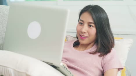 Young-business-freelance-Asian-woman-working-on-laptop-checking-social-media-while-lying-on-the-sofa-when-relax-in-living-room-at-home.-Lifestyle-latin-and-hispanic-ethnicity-women-at-house-concept.