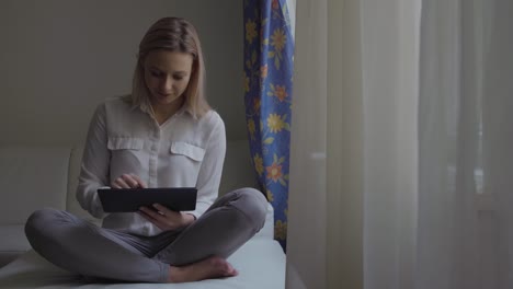 Eine-junge-schöne-Frau-sitzt-querbeinig-auf-einer-Couch-in-einer-Wohnung-und-arbeitet-auf-einem-Tablet