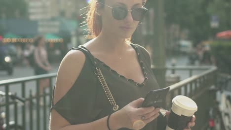 Joven-atractiva-mujer-escribiendo-un-mensaje-de-texto-en-su-teléfono-inteligente-en-la-salida-del-metro-en-la-calle,-sosteniendo-su-café-en-la-otra-mano,-durante-la-tarde-soleada-de-verano-en-París.-De-moda-y-fresco,-cámara-lenta.