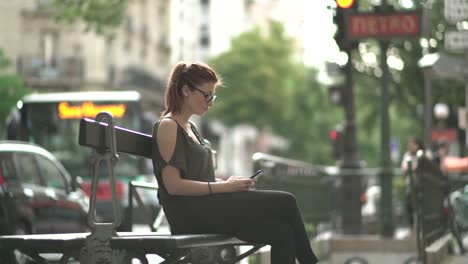 Attractive-redhead-woman-with-glasses,-freckles,-piercings-and-red-hair-writing-a-text-message-on-her-smartphone-sitting-on-street-bench,-during-sunny-summer-in-Paris.-Tilt-up-4K-UHD.-Trendy.