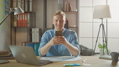 Creative-Entrepreneur-Sitting-at-His-Desk-Works-on-a-Laptop-in-the-Stylish-Office,-Uses-Smartphone-for-Social-Media-Apps,-Emailing-Business-Associates,-Reading-News,-Browsing-through-Internet