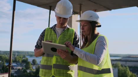 Trabajadorde-la-construcción-hombre-y-arquitecto-mujer-en-un-casco,-discutir-el-plan-de-construcción-de-la-casa,-hablarse-unos-a-otros-sobre-el-diseño,-sosteniendo-una-tableta,-mirar-los-dibujos,-fondo-de-los-rayos-del-sol