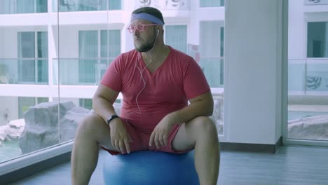 Funny-fat-male-in-pink-glasses-and-in-a-pink-t-shirt-is-engaged-on-a-fit-ball-in-the-gym-depicting-a-girl.-A-wonderful-man-listens-to-music-and-dances-on-the-ball-in-the-gym