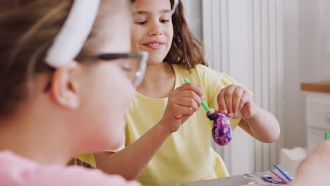 Dos-chicas-sentadas-alrededor-de-la-mesa-de-la-cocina-con-orejas-de-conejo-decorando-huevos-para-Semana-Santa---tiro-en-cámara-lenta