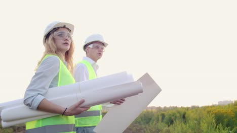 Ein-Mann-und-eine-Baufrau-mit-Zeichnungen-in-weißen-Helmen-gehen-und-reden-auf-der-Baustelle