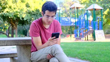 Junger-Mann-mit-seinem-Smartphone-sitzt-in-einer-Parkbank