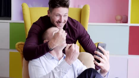 Gay-couple-using-phone-on-arm-chair-together.-Showing-ring-via-video-call.