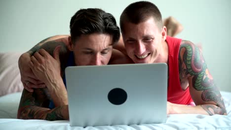 Pareja-gay-en-la-cama-usando-computadora-portátil.-Abrazando-y-sonriendo.