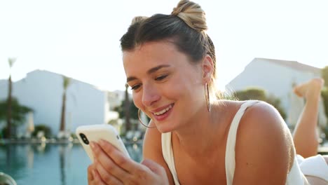 Close-up-of-the-pretty-Caucasian-young-girl-sunbathing-at-the-swimming-pool-and-using-her-smartphone-for-playing-or-social-media,-then-smiling-happily-to-the-camera.-Portrait.