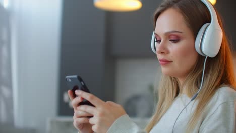 Young-girl-listening-to-music-in-headphones-looking-at-the-screen-of-the-smartphone.-Relax-at-home