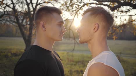 Close-up-of-two-Caucasian-sportsmen-looking-at-each-other-in-sunlight.-Serious-adult-boys-standing-outdoors.