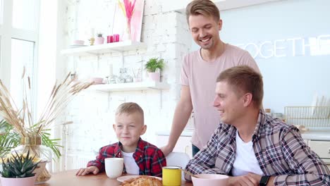 Glückliche-schwule-Familie-zwei-Väter-und-Sohn-frühstücken-in-der-Küche.