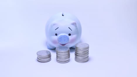 Light-blue-piggy-bank-with-many-money-coins-on-white-background-and-money-saving-ideas.-Timelapse-4K