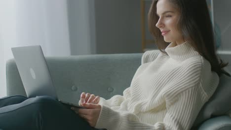 Beautiful-Young-Woman-Works-on-Laptop-Computer-while-Sitting-on-the-Chair.-Sensual-Girl-Wearing-Sweater-Works-On-Notebook;-Studies,-Surfs-Internet,-Uses-Social-Media-while-Relaxing-in-Cozy-Apartment