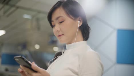 Retrato-de-mujer-asiática-con-auriculares-escuchando-música-y-usando-teléfono-inteligente-para-chatear-con-amigos-o-navegar-mientras-espera-un-tren.-Tecnología-en-la-vida-cotidiana-y-los-viajes.