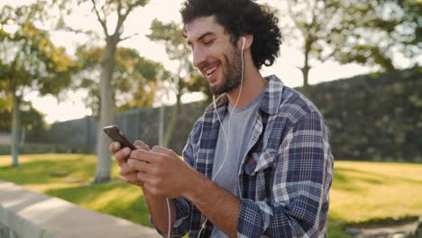 Primer-plano-de-un-joven-feliz-sonriente-con-auriculares-blancos-en-los-oídos-usando-el-móvil-para-mensajes-de-texto-en-el-parque