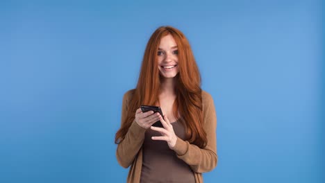 Redheaded-girl-in-brown-t-shirt-and-cardigan.-She-is-using-smartphone,-smiling-and-looking-surprised-while-posing-against-blue-background.-Close-up