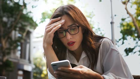 Business-woman-in-glasses,-white-shirt.-She-feeling-disappointed-while-reading-news-using-smartphone,-sitting-in-outdoor-cafe.-Close-up,-slow-motion