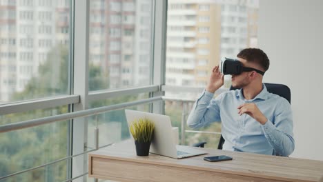 A-young-man-sitting-at-a-desk-in-the-office-uses-augmented-reality-glasses-to-work-on-business-projects-in-various-fields.