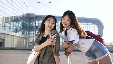 Charming-cheerful-25-years-old-asian-female-travelers-standing-near-modern-airport-and-using-their-smartphones