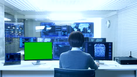 Back-View-of-the-IT-Technician-Working-on-with-Neural-Network-on-His-Personal-Computer-with-Green-Screen-Mock-up.-He-Works-in-a-Big-System-Control-Data-Center-with-Multiple-Monitors-Showing-Graphics.