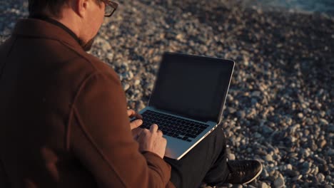 Man-is-working-with-a-laptop-sit-on-pebble-shore-of-sea-in-evening