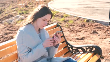 Portrait-of-beautiful-brunette-reading-and-typing-a-message-in-her-phone-sitting-on-the-bench-in-park.