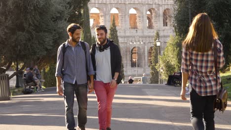 Young-happy-gay-couple-tourists-walk-in-park-road-with-trees-colosseum-in-background-in-rome-at-sunset-holding-hands-gelousy-when-beautiful-attractive-girl-walks-by-glancing-lovely-slow-motion-colle-oppio