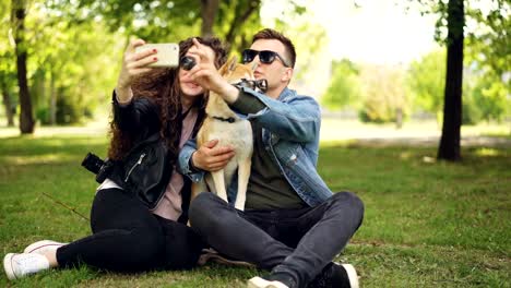 Mujer-bonita-está-tomando-selfie-con-su-novio-y-adorable-perro-con-smartphone-mientras-descansa-en-el-parque-en-el-césped.-Los-seres-humanos-y-los-animales-llevan-gafas-de-sol.