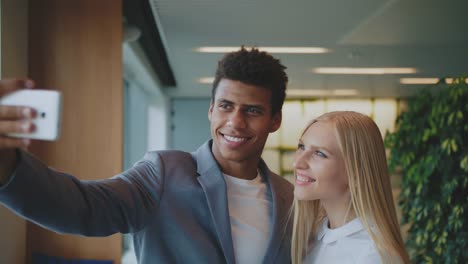 Lachen-Sie-diverse-Kollegen-nehmen-Selfie-im-Amt.-Fröhlich-schwarzer-Mann-mit-lachen-blonde-Frau-unter-Selfie-mit-Smartphone-in-modernen-Büros-mit-Spaß