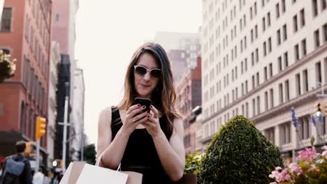 Happy-attractive-Caucasian-businesswoman-with-shopping-bags-chatting-with-friends-on-smartphone-social-app-in-New-York