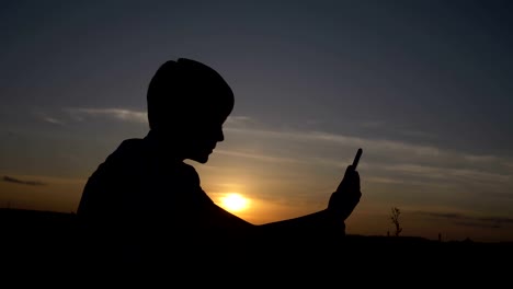 Silhouette-eines-jungen-verwendet-eine-Tablette-bei-Sonnenuntergang-im-Feld-liest-etwas-auf-dem-Tablet-im-freien