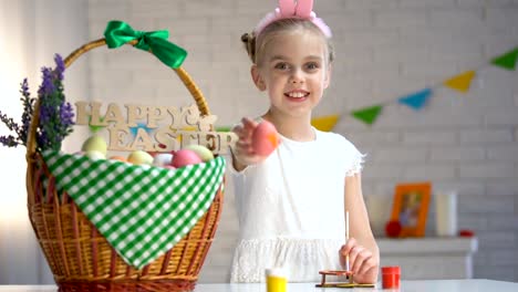 Creative-girl-in-white-dress-showing-painted-Easter-egg,-decorated-basket-table