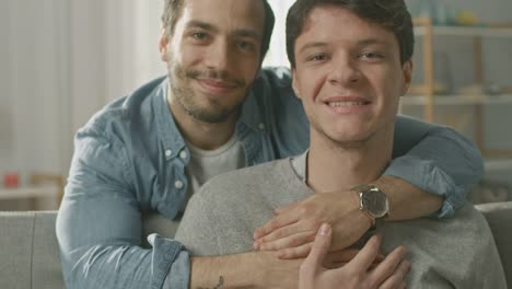 Portrait-of-a-sanfte-männliche-Gay-Couple-zu-Hause.-Junger-Mann-sitzt-auf-einer-Couch,-seinem-Partner-umarmt-ihn-von-hinten.-Sie-sind-glücklich-und-Lächeln.-Zimmer-ist-Modern-eingerichtet.