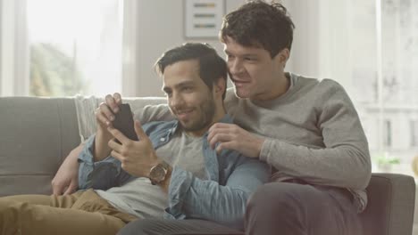 Süße-männliche-Gay-paar-verbringen-Zeit-zu-Hause.-Sie-sind-unten-auf-dem-Sofa-liegen-und-ein-Smartphone-benutzen.-Stöbern-sie-Online.-Partners-Hand-ist-um-seine-Geliebte.-Sie-Lächeln-und-lachen.-Zimmer-ist-Modern-eingerichtet.