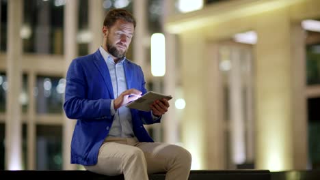 Concentrated-middle-aged-man-sitting-on-bench-outdoors-in-evening-and-browsing-Internet-on-tablet-computer-via-wifi