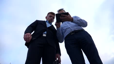 Low-angle-view-of-young-businessman-showing-a-presentation-on-screen-of-tablet-pc-to-his-colleague-in-city.-Successful-entrepreneurs-working-on-digital-tablet.-Blue-sky-at-background.-Slow-motion