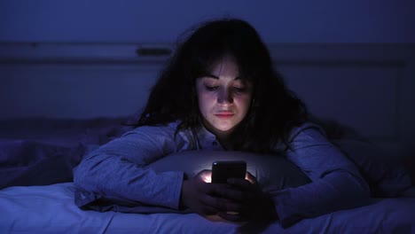 Video-4K-de-una-joven-adicta-charlando-y-navegando-por-Internet-usando-su-teléfono-inteligente-soñoliento,-aburrido-y-cansado-tarde-por-la-noche.-Luz-oscura-dramática.-En-Internet,-adicción-móvil-y-el-concepto-de-insomnio.