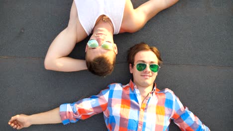 Top-view-of-young-happy-male-couple-in-sunglasses-lying-on-rooftop-of-high-rise-building-and-smiling.-Handsome-gay-boys-looking-at-camera-and-enjoying-to-spend-time-on-roof.-Close-up-Slow-motion