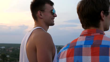 Dolly-shot-of-young-male-couple-in-sunglasses-standing-on-the-edge-of-rooftop-and-talking.-Handsome-gay-boys-rest-on-roof-of-high-rise-building-and-enjoying-beautiful-cityscape.-Close-up-Slow-motion