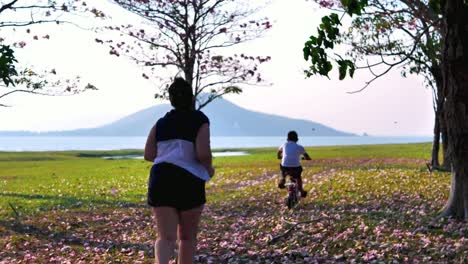 An-Asian-woman-jogging-in-natural-sunlight-in-the-evening,-along-with-his-son-riding-a-bicycle.--exercising-for-good-health.-Slow-Motion