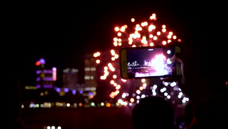 Human-hand-holding-smartphone-filming-a-video-of-night-fireworks,-people-and-buildings-in-the-background-slow-motion.