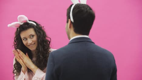 A-young-beautiful-girl-stands-on-a-pink-background-and-looks-at-the-boy-who-is-standing-back-to-the-camera.-A-gray-skirt-and-a-pink-sweater-with-hooked-ears-on-the-head.-Smiling-simulates-the-movements-of-the-hand-in-hand.-Easter-concept.